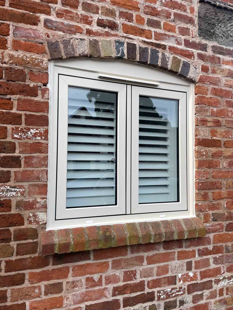 A white uPVC double-glazed window with internal blinds, set in a red brick wall with an arched brick detail above.