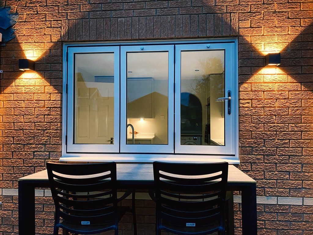 Blue-framed window on brick wall with outdoor table and chairs, illuminated by wall lights at night.