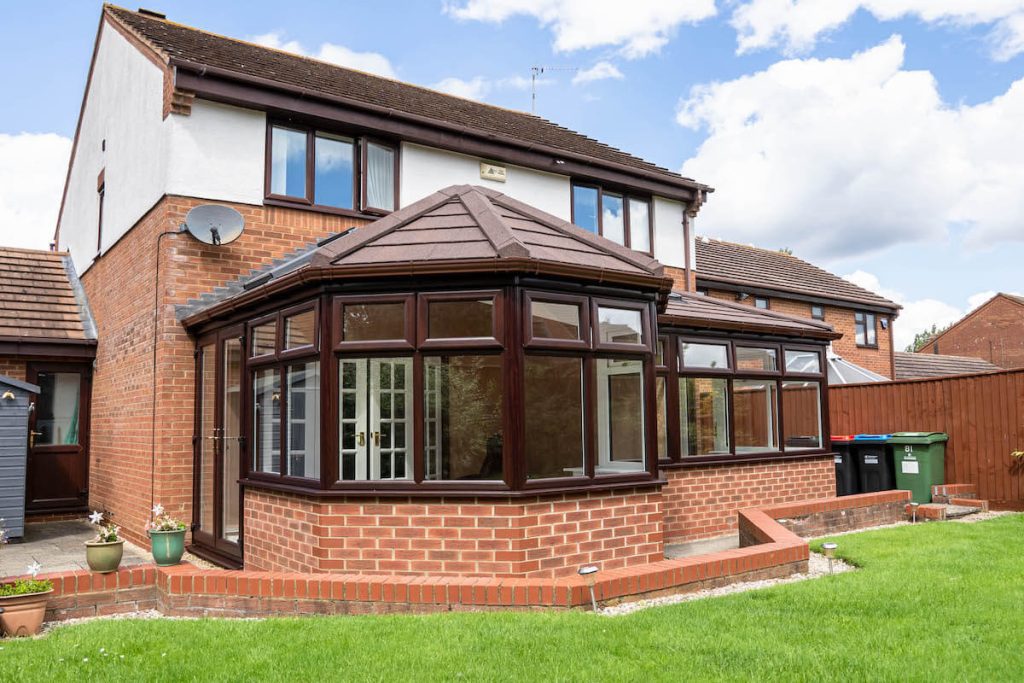 A semi-detached UK home with a brown conservatory featuring a tiled roof and uPVC frames, surrounded by a garden. The image reflects the page's focus on modern conservatory designs and installations.