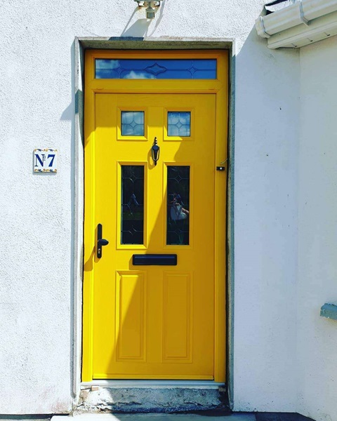 A bright yellow composite door with two glass panels and a decorative transom window, set against a white exterior.