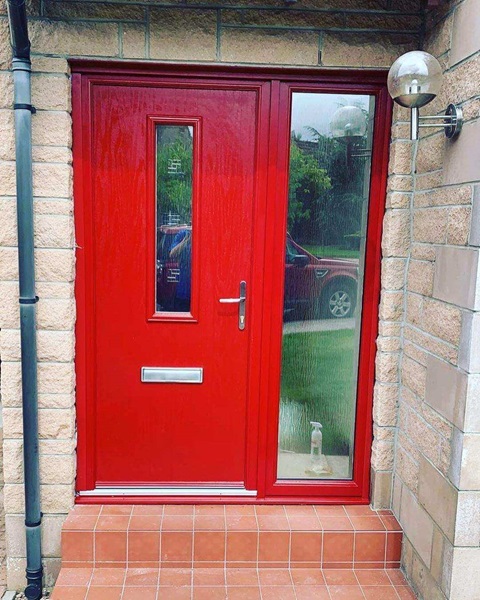 A bright red composite door featuring a slim vertical glass panel and a large frosted glass side panel, set against a brick façade.