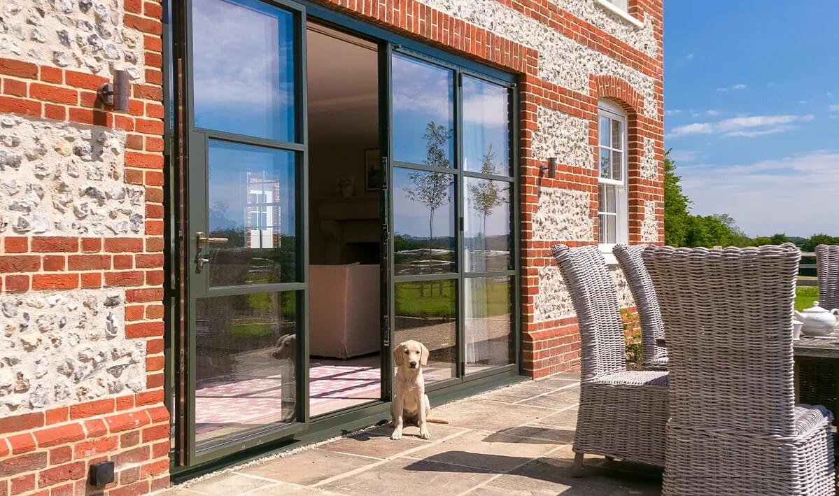 Green Origin bifold door on a stone cottage, leading to a cosy interior, with a dog sitting by the doorway and a patio setup outside.